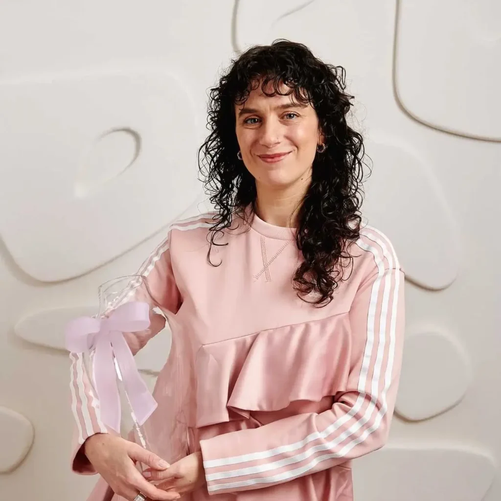 A woman with curly dark hair wearing a pink top stands in front of a textured white wall, holding a clear trophy with a light purple bow—celebrating her creative design at the Linda Pro concept store.